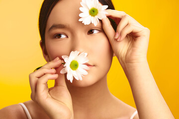 Asian woman is captured in a closeup, holding a flower near her face against a bright yellow background. She smiles softly, exuding calm, beauty, and a fresh, natural mood.