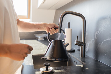 Close-up of man pouring water into black kettle from stylish faucet in modern kitchen. Concept of daily routine, home lifestyle, and cooking preparation. Symbol of household comfort, minimalistic