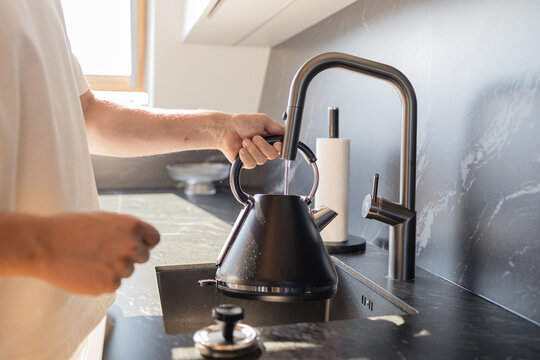 Close-up of man pouring water into black kettle from stylish faucet in modern kitchen. Concept of daily routine, home lifestyle, and cooking preparation. Symbol of household comfort, minimalistic