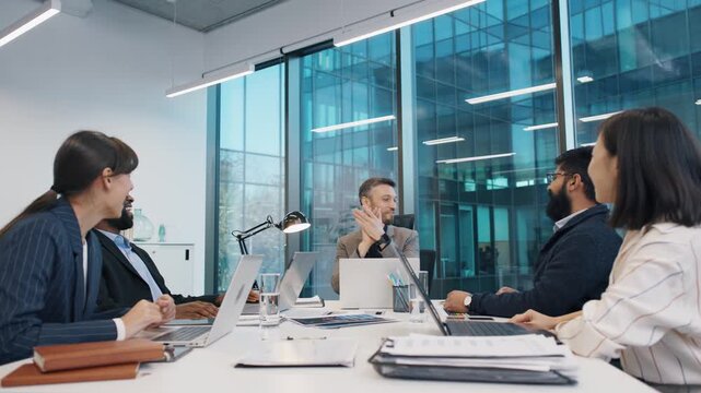 Business team laughing together during relaxed strategy session in modern glass office, led by man in grey blazer. Diverse professionals seated around laptops and charts, exchanging ideas.