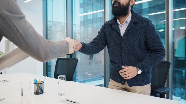 South Asian man with beard attentively answering questions during job interview with Caucasian man holding folder. Business setting with glass walls and professional tone. Males shaking hands.