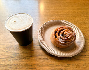 Coffee latte art on wood table with a bun with raisins in cafe close-up. Cappuccino with a pattern in a paper cup and bun on a wooden table in a coffee shop top view. The concept of food and drinks.