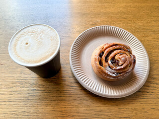 Coffee latte art on wood table with a bun with raisins in cafe close-up. Cappuccino with a pattern in a paper cup and bun on a wooden table in a coffee shop top view. The concept of food and drinks.
