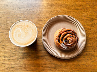 Coffee latte art on wood table with a bun with raisins in cafe close-up. Cappuccino with a pattern in a paper cup and bun on a wooden table in a coffee shop top view. The concept of food and drinks.