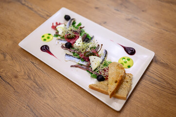 Fine dining seafood starter with sardines, cheese, olives, sprouts, and toast on a white plate with artistic sauces on rustic wooden restaurant table.