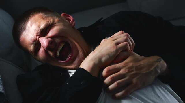A young man in a black shirt lies on the couch, clutching his chest with his hands from severe pain in his heart.
The man is having a heart attack and panic attack.
The man has a heart condition.
