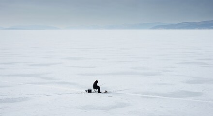 A lone person sits on a small chair, actively ice fishing on a vast, frozen, snow-covered lake under a grey, overcast sky with distant mountains.