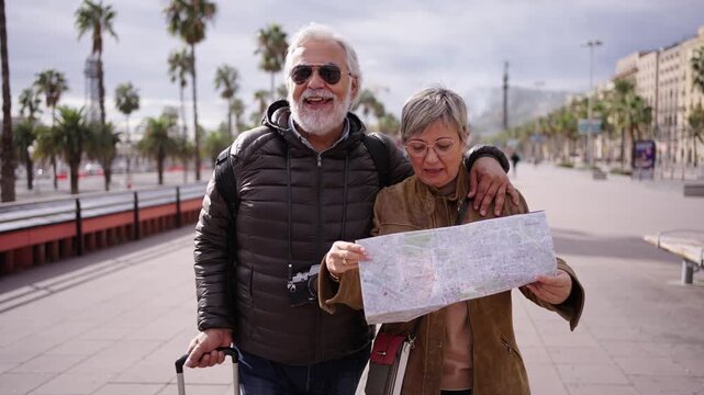 Senior Caucasian tourist couple walking with suitcase together pointing enjoying holidays. Grey hair pair laughing having fun sightseeing in European city center. Happy adults strolling holding map