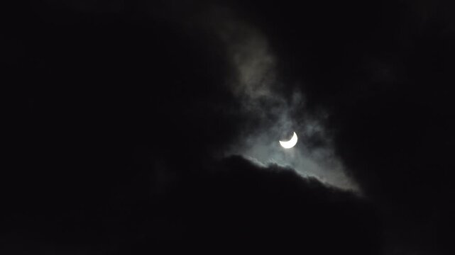 A partial solar eclipse glows while it's seen through dark clouds moving across the sky.