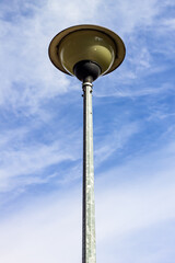 Tall, modern street lamp with a light-green hue stands against a bright blue sky with wispy clouds, captured from a low-angle perspective, emphasizing its verticality and urban design.

