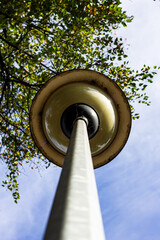 Extreme low-angle view of a vintage yellow street lamp and pole stretching into a bright blue sky, with green tree leaves framing the top, showcasing nature and urban contrast.

