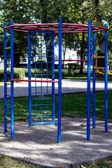 Brightly colored (blue and red) metal monkey bars and fitness frame stand empty in an outdoor park, symbolizing urban fitness, play, and vibrant equipment against a green background.

