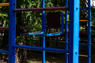Vibrant blue metal monkey bars dominate the foreground of an outdoor playground or street workout area, with lush green trees in soft focus behind, creating a dynamic pop of color and depth.

