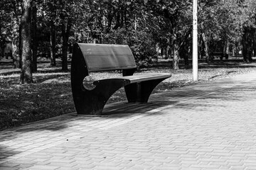 Modern, uniquely shaped park bench sits empty on a brick walkway in a black and white park setting, emphasizing solitude and design.

