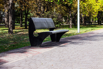 A modern, stylish black bench sits empty on a brick-paved path in a sunlit park. The scene captures the tranquility of early autumn and a peaceful rest area.

