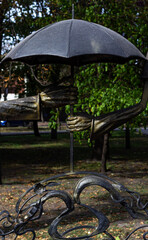A charming bronze sculpture of two hands holding an umbrella, symbolizing love or care, set against a blurred park background. Focus on art, romance, and urban decor.


