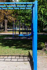 A bright blue monkey bar frame at a park playground on a sunny day. The metal structure casts a sharp grid shadow onto the ground, framed by green grass and trees.


