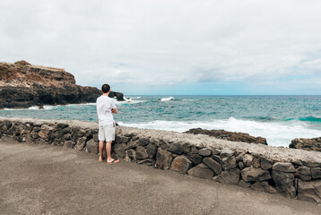 A brunette tourist watches the waves crashing against the rocks.
