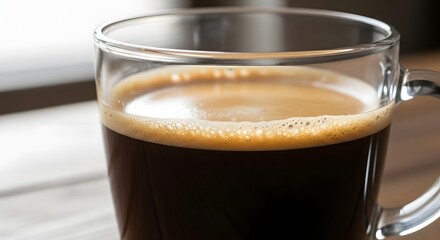 A close-up of a clear glass mug filled with black coffee and crema on a wooden table.