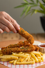 Hand holding breaded chicken stick with fries and sauce