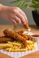 Hand dipping breaded chicken stick in sauce with fries