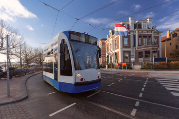 Naklejka premium Blue and white tram passing by historic buildings and Dutch flag in Amsterdam Netherlands
