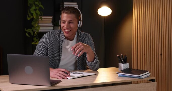 Man in headphones taking notes while passing online test via laptop at table indoors