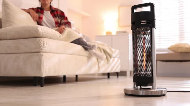 Woman with blanket sitting on sofa at home, focus on electric heater
