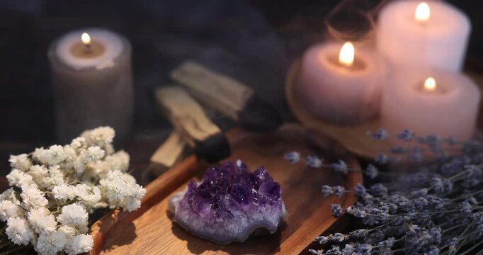 Beautiful amethyst gemstone, flowers, palo santo sticks and burning candles on table in smoke, closeup