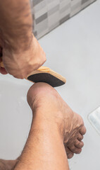 Close-up of a male person using a wooden foot file to exfoliate dry skin on their heel in a bright bathroom, showcasing personal care and hygiene practices for healthy feet
