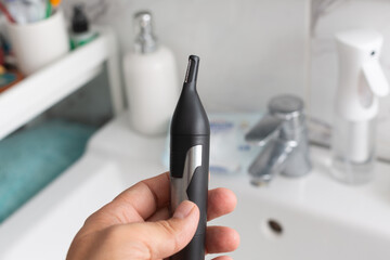 Hand holding a black electric nose hair trimmer in a modern bathroom setting with sleek fixtures and personal care items, showcasing grooming and hygiene essentials