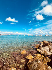 Clear turquoise water gently laps against rocky shoreline under a bright blue sky with fluffy clouds, creating a serene coastal landscape perfect for relaxation and escape