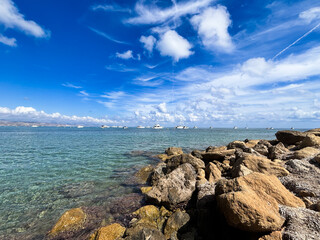 Scenic coastal view featuring clear turquoise waters, rocky shoreline, and boats in the distance under a bright blue sky with fluffy white clouds, evoking tranquility and natural beauty