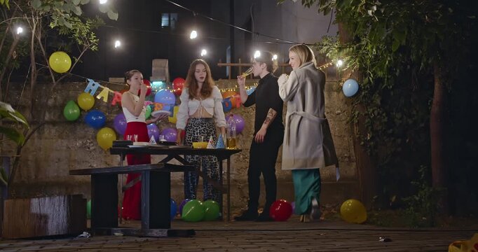 Friends gather in a backyard at night for a birthday celebration with cake, drinks, balloons, and string lights.