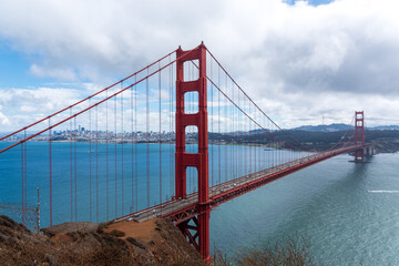 The Golden Gate Bridge stretching across San Francisco Bay, with the city skyline visible in the distance under a mix of clouds and blue sky.
