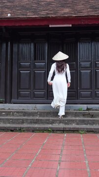 
4k Vertical video.Woman in traditional Vietnamese &aacute;o d&agrave;i and conical hat walking in front of ancient wooden door, symbol of Vietnam culture and travel lifestyle.