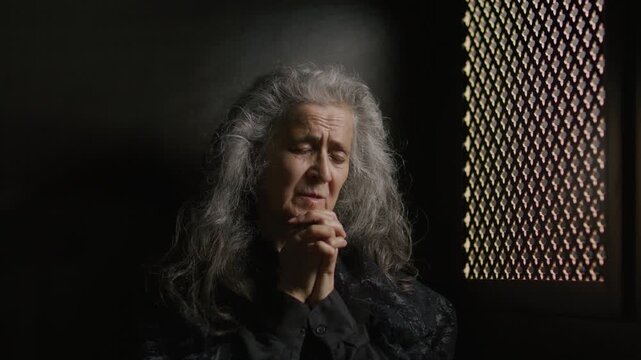 Sorrowful elderly woman with long gray hair clasping hands and praying to God while sitting alone inside confessional booth in church