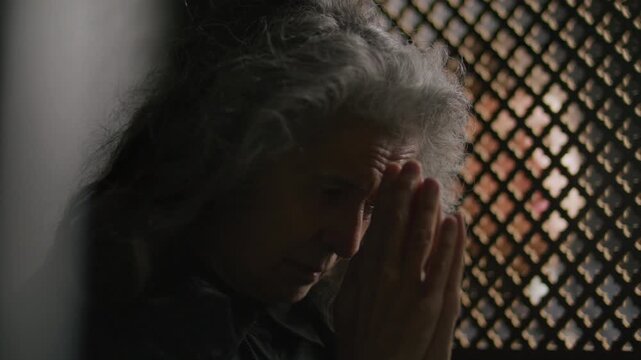 Senior woman with long grey hair clasping hands in prayer as listening to priest talking through lattice screen of confessional booth
