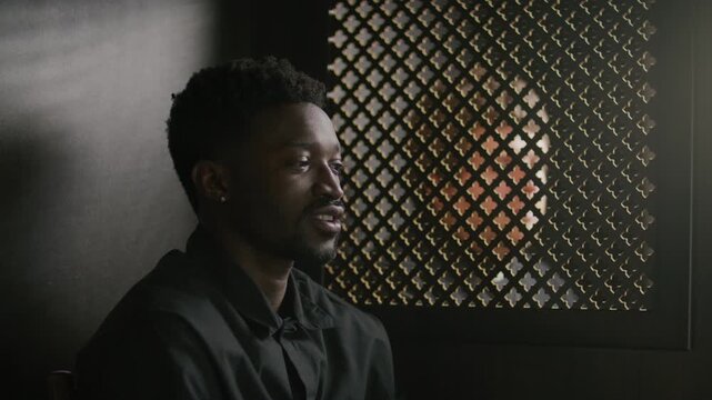 Young Black parishioner sitting in confessional booth, talking emotionally with priest through lattice screen
