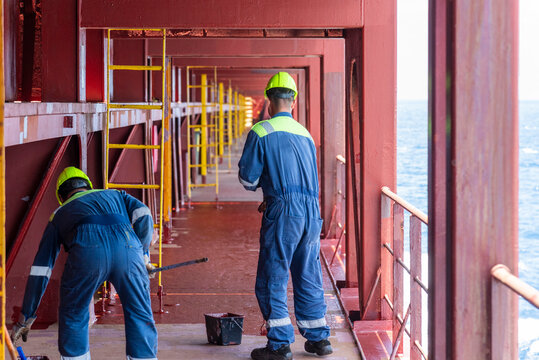 Two male crewmembers in blue coveralls and yellow hard hats are painting the deck of a large container ship - real workplace.