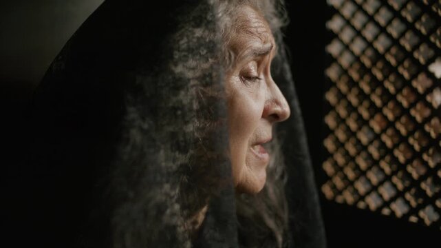 Elderly woman in black mantilla speaking quietly to priest through lattice screen during spiritual confession in church