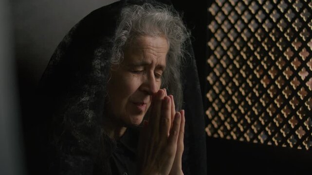 Elderly woman with gray hair wearing black lace veil confessing her sins and praying with her hands clasped together during conversation with priest through lattice screen in church