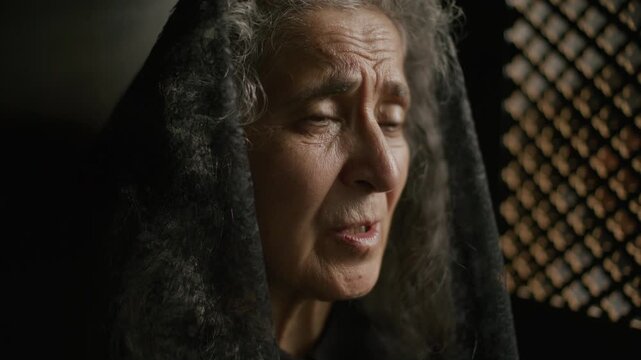 Elderly woman in black lace veil talking emotionally and whispering prayer with closed eyes and clasped hands near wooden lattice screen in confessional booth
