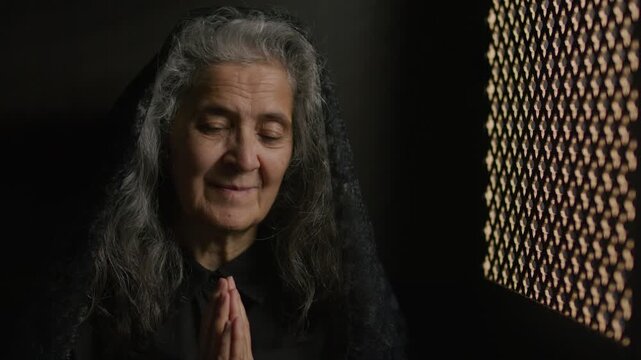 Senior woman with long grey hair, wearing black mantilla and holding her hands in prayer as making confession to priest through lattice screen