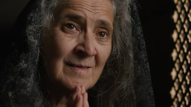 Senior grey-haired woman in lace veil clasping hands beneath chin as speaking with priest through perforated screen during spiritual confession. Close-up view