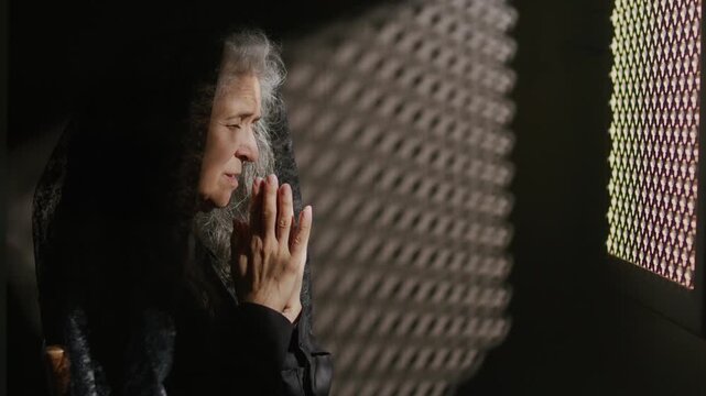 Elderly woman in black mantilla bowing her head and holding hands together in prayer near confessional screen in church. Profile view