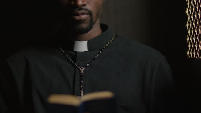 Black priest wearing clerical collar and rosary reading Bible near lattice partition inside confessional booth. Close-up shot
