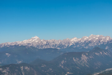 The snow-capped Julian Alps, with Mount Triglav, majestically dominate the horizon under a clear blue sky, viewed from Weinhasch in the Karawanken, highlighting Slovenia's pristine alpine grandeur.