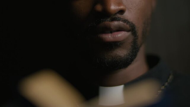 Close-up view of African American priest in clerical collar reading from bible in dimly lit confessional booth