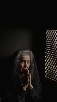 Senior grey-haired woman in black mantilla holding rosary and praying with hands clasped while sitting in confessional booth. Vertical format footage